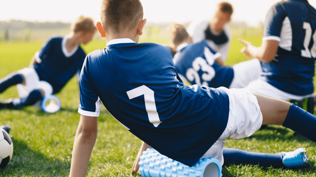 Młody piłkarz rolujący mięśnie nóg na wałku piankowym w ramach regeneracji po treningu.